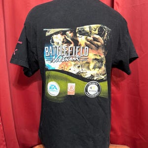 May include: Black t-shirt featuring a Battlefield Vietnam graphic on the back. The design includes a soldier, explosions, and the game's logo in blue and white. The shirt also has EA Games and NVIDIA logos.