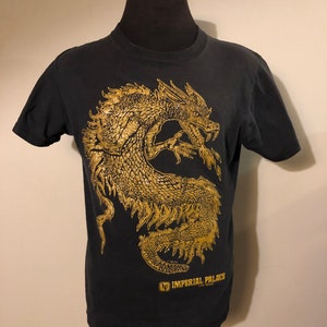 以下が含まれることがあります： 前面に金の龍のグラフィックと「インペリアルパレスラスベガス」の文字がプリントされた黒いTシャツです。