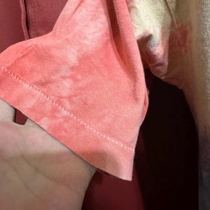 K&ouml;nnte beinhalten: Nahaufnahme des &Auml;rmels eines Batik-Shirts. Der &Auml;rmel ist hellrosa mit einem wei&szlig;en und gelben Batik-Muster. Der &Auml;rmel ist umgeschlagen und die N&auml;hte sind sichtbar.