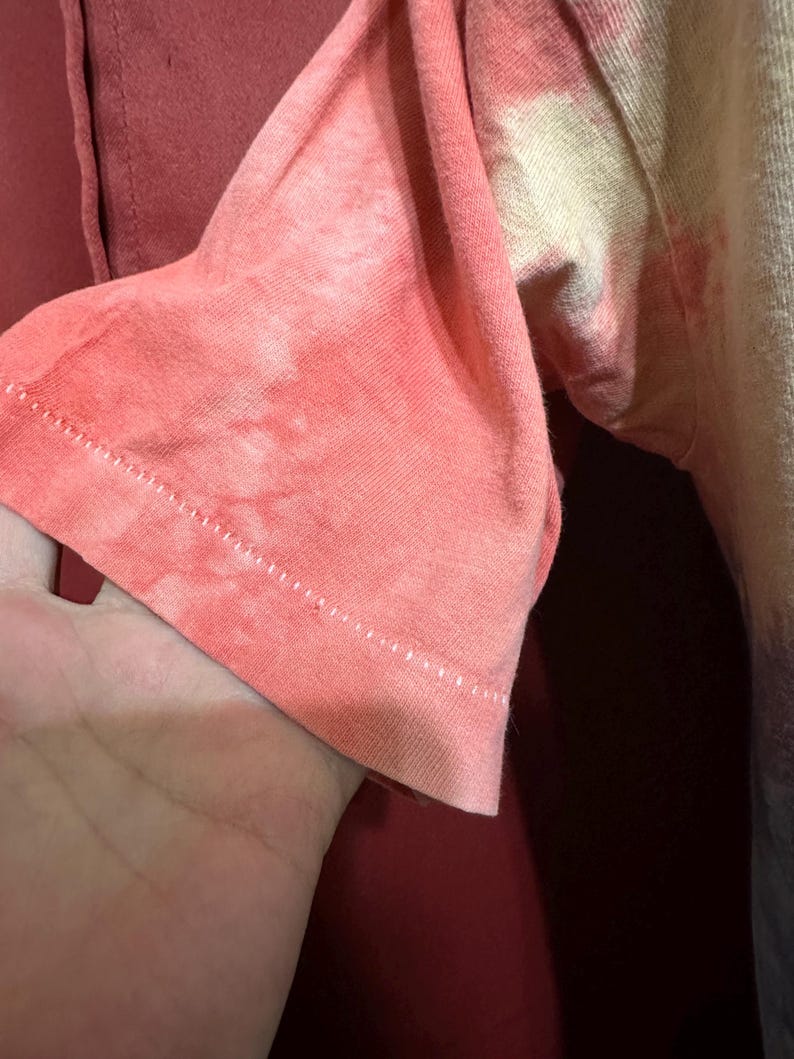 K&ouml;nnte beinhalten: Nahaufnahme des &Auml;rmels eines Batik-Shirts. Der &Auml;rmel ist hellrosa mit einem wei&szlig;en und gelben Batik-Muster. Der &Auml;rmel ist umgeschlagen und die N&auml;hte sind sichtbar.