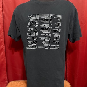 以下が含まれることがあります： 白いテキストデザインの黒いTシャツ。テキストは、コンピューターコマンドまたはコードのリストのようです。シャツはマネキンに表示されています。背景は赤いカーテンです。