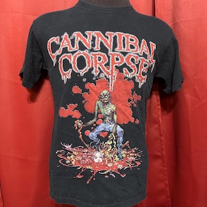 Puede incluir: Camiseta negra con un estampado gráfico de un zombi de pie en un charco de sangre. El texto "Cannibal Corpse" está impreso en rojo y blanco sobre el zombi.