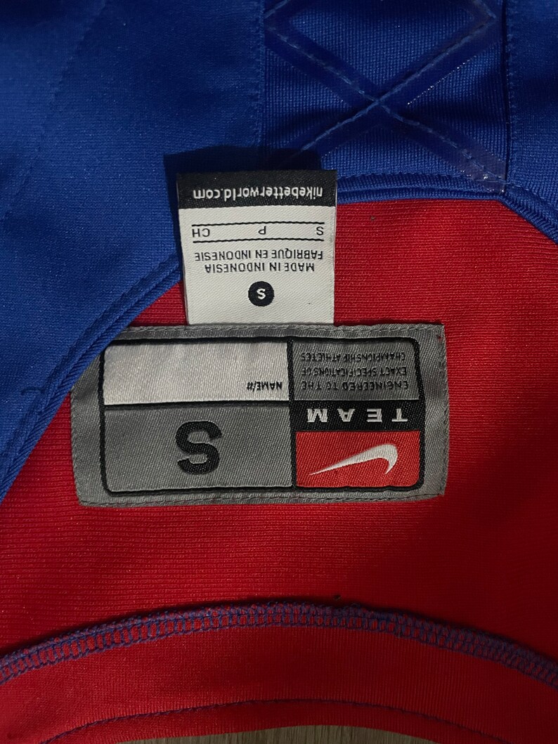 May include: A gray and white tag with the text "TEAM ENGINEERED TO THE NAME/# EXACT SPECIFICATIONS OF CHAMPIONSHIP ATHLETES" and a Nike swoosh logo. The tag also has the text "S" in a larger font.