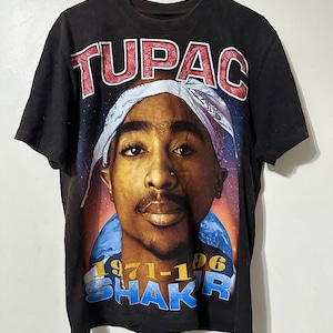May include: Black t-shirt featuring a portrait of Tupac Shakur with the word "TUPAC" in red block letters above the image. The shirt also includes the text "1971-1996 SHAKUR" in blue and white.
