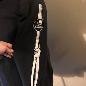 May include: Black hooded sweatshirt with a white skeleton arm graphic on the sleeve. The graphic includes a skull with the word "SKATE" written on a banner below it.
