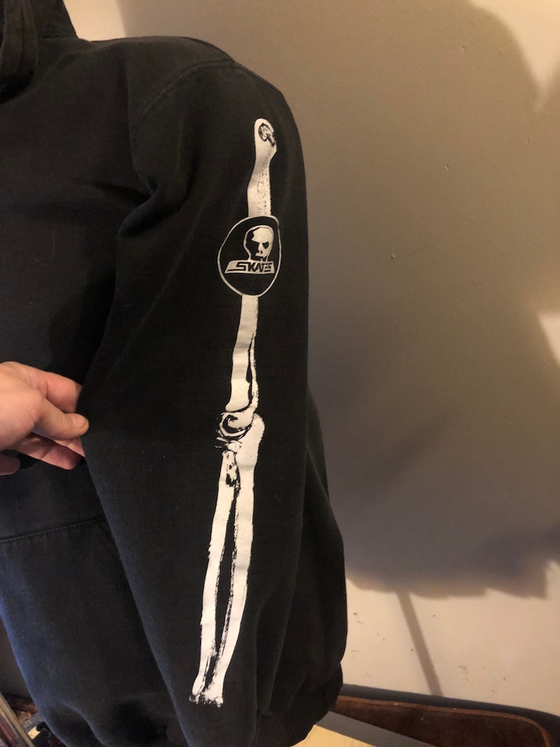 May include: Black hooded sweatshirt with a white skeleton arm graphic on the sleeve. The graphic includes a skull with the word "SKATE" written on a banner below it.