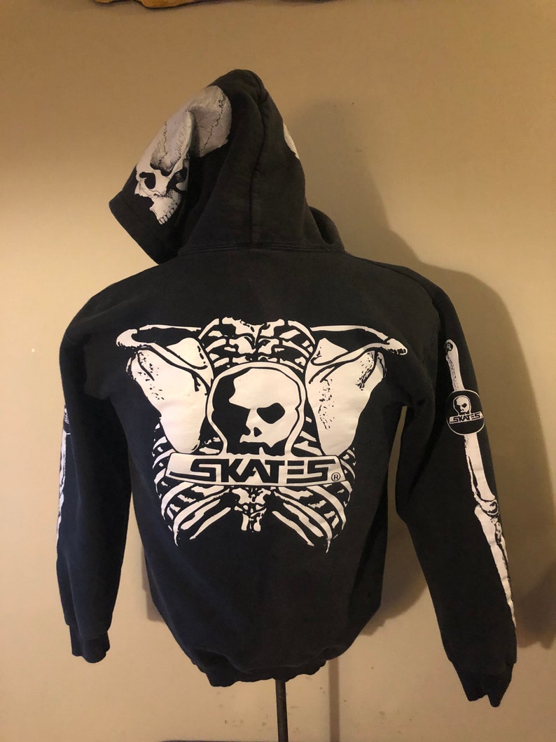 May include: Black zip-up hoodie with a white skeleton graphic on the back. The graphic includes the text "SKATES" and a skull. The hoodie has a white skull graphic on the hood.