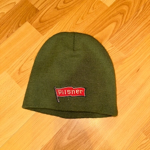 Puede incluir: Un gorro de punto verde con una bandera bordada en rojo que dice "Pilsner".