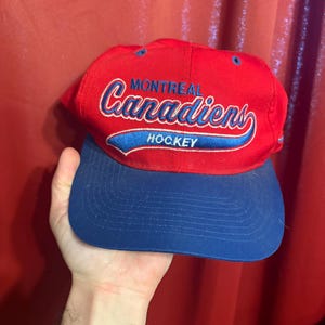 May include: A red and blue baseball cap featuring the text "Montreal Canadiens Hockey" in white and blue script. The cap has a blue brim and a red crown. The hat is a sports-themed accessory.