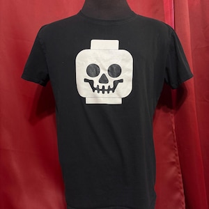 May include: A black short-sleeved t-shirt featuring a large white graphic on the chest. The graphic depicts a stylised toy minifigure head silhouette with a black skull face design, including hollow eye sockets, a triangular nose, and teeth.