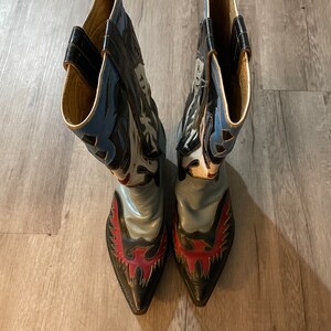 May include: A pair of black cowboy boots with blue, white, and red leather designs. The boots have pointed toes and a decorative eagle design on the back.