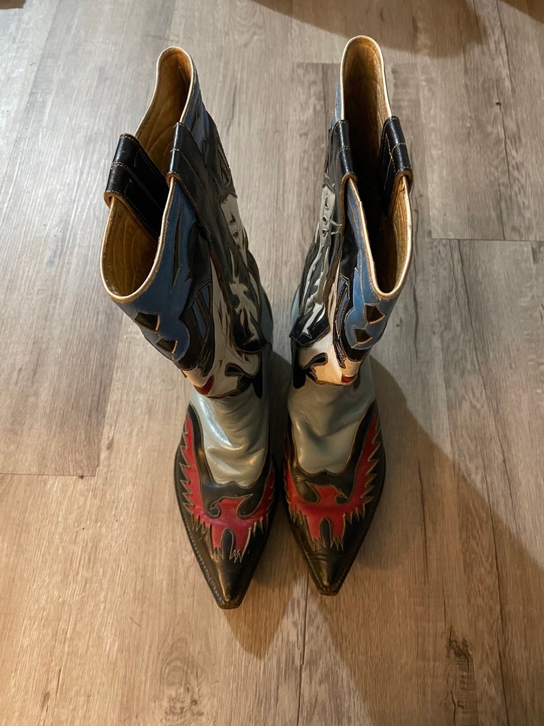May include: A pair of black cowboy boots with blue, white, and red leather designs. The boots have pointed toes and a decorative eagle design on the back.