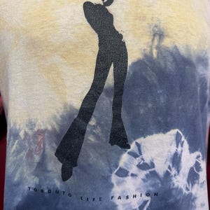 K&ouml;nnte beinhalten: Ein Batik-T-Shirt mit einer schwarzen Silhouette einer Frau in einer stilvollen Pose. Das Shirt tr&auml;gt den Text "Toronto Life Fashion" auf der Unterseite.