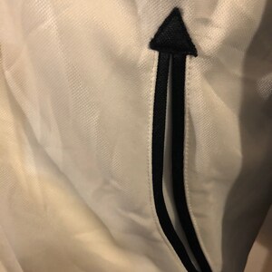 May include: White athletic jacket with a black zipper detail and a black arrow pointing upwards on the pocket.