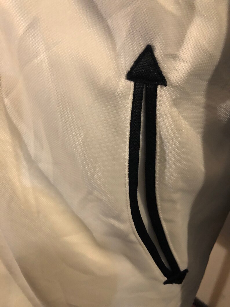 May include: White athletic jacket with a black zipper detail and a black arrow pointing upwards on the pocket.