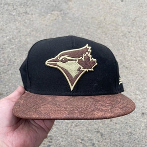 Könnte beinhalten: Eine schwarze Baseballkappe mit braunem Leder-Schirm. Die Kappe hat ein golden gesticktes Logo eines Blauhähers mit einem Ahornblatt auf dem Kopf.