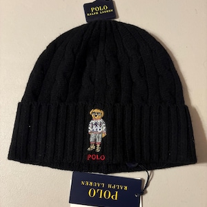 May include: Black knit beanie with a cable knit design and a folded brim. Features an embroidered bear wearing sunglasses and a sweater. The word "POLO" is embroidered in red below the bear. Includes a hang tag that says "POLO RALPH LAUREN".