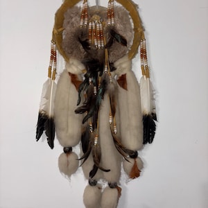 Puede incluir: Un atrapasueños artesanal con un marco circular de color beige y un diseño central de piel. Cuenta con plumas blancas y marrones, hebras de cuentas y elementos de lana suave blanquecina. El atrapasueños está suspendido sobre un fondo blanco.