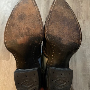 May include: A pair of brown leather cowboy boots with a white and black design. The boots have a pointed toe and a rubber sole with the text "Justin Boots" on the bottom.