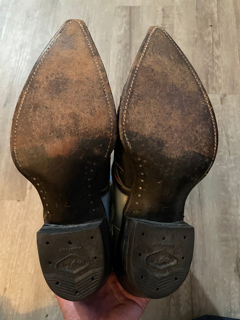 May include: A pair of brown leather cowboy boots with a white and black design. The boots have a pointed toe and a rubber sole with the text "Justin Boots" on the bottom.