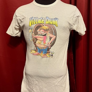 May include: White t-shirt with a cartoon graphic of a man holding a beer mug, with the text "VILLAGE DRUNK" above the image. The man has a long tongue and is standing next to a beer barrel. The shirt is displayed on a mannequin.