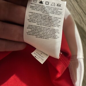 May include: White care tag with black text and symbols. The text reads "Machine wash cold/Wash with like colors immediately/Do not allow to lay on itself/Do not use softeners/Remove when wet/Do not bleach/Tumble dry low/Cool iron/Do not iron design/Do not dry clean".