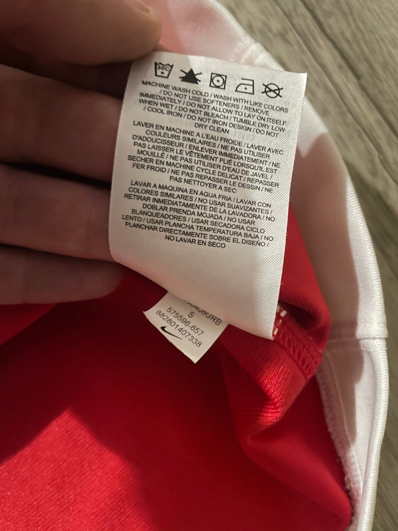 May include: White care tag with black text and symbols. The text reads "Machine wash cold/Wash with like colors immediately/Do not allow to lay on itself/Do not use softeners/Remove when wet/Do not bleach/Tumble dry low/Cool iron/Do not iron design/Do not dry clean".