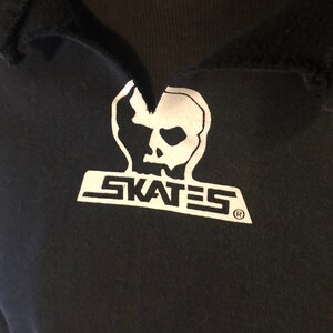 May include: Black t-shirt with a white skull graphic and the word "SKATES" printed below it. The graphic is stylized with a jagged edge and a registered trademark symbol.