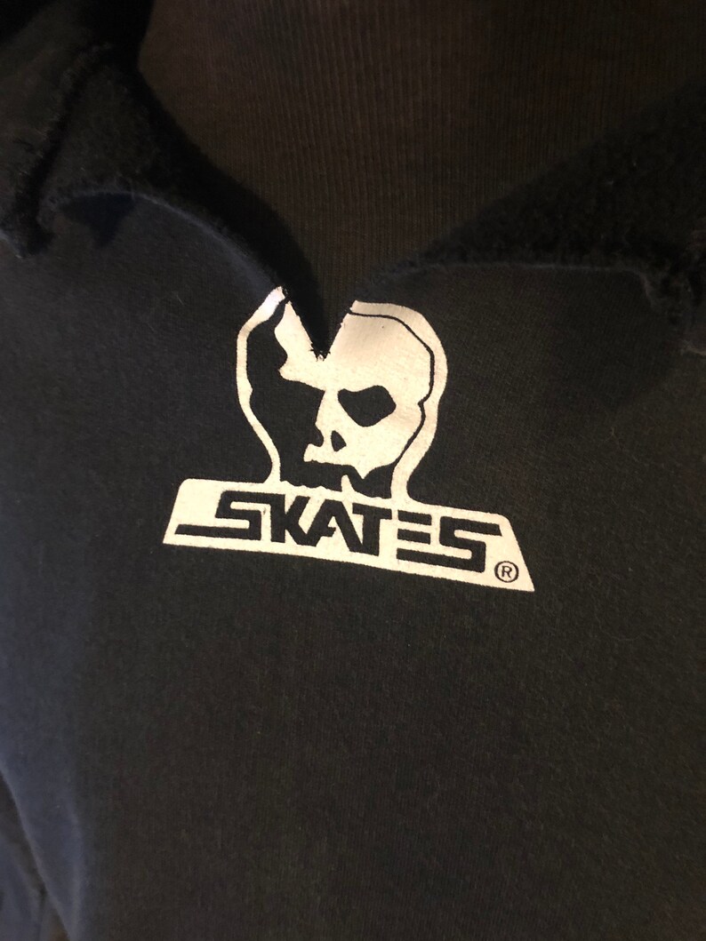 May include: Black t-shirt with a white skull graphic and the word "SKATES" printed below it. The graphic is stylized with a jagged edge and a registered trademark symbol.