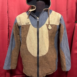 May include: A hooded jacket with a patchwork design in shades of brown, tan, and blue. The jacket has a full-length zipper and a black interior lining. The hood is brown with a black interior. The jacket is displayed on a mannequin.