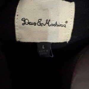 May include: Close-up of a white label with the text "Deus Ex Machina" in cursive. Below the brand name is a smaller label with the size "L" and the text "MADE IN CHINA". The background is black.