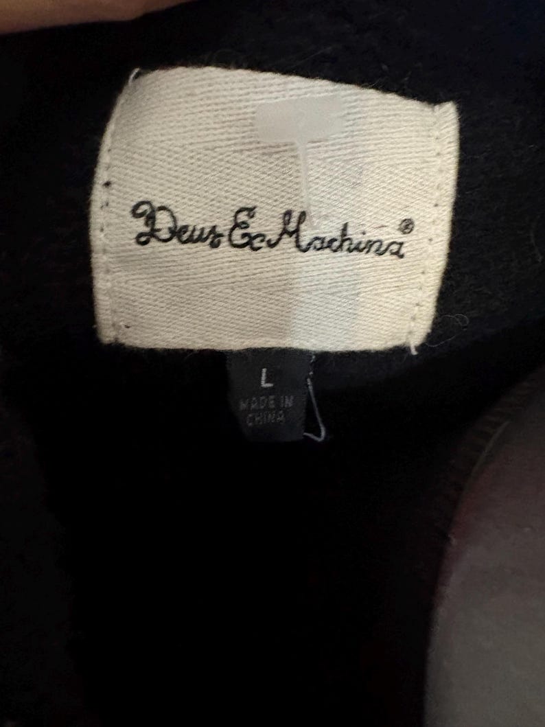 May include: Close-up of a white label with the text "Deus Ex Machina" in cursive. Below the brand name is a smaller label with the size "L" and the text "MADE IN CHINA". The background is black.
