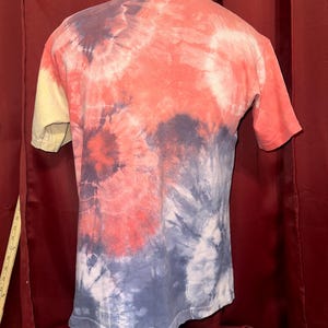 K&ouml;nnte beinhalten: Ein T-Shirt im Tie-Dye-Look mit gelber, orangefarbener, blauer und wei&szlig;er Farbgebung. Das Shirt befindet sich auf einem Mannequin.