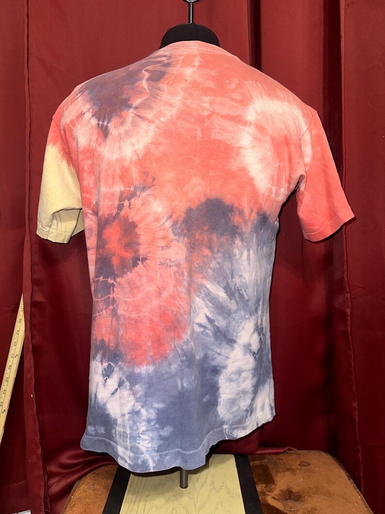 K&ouml;nnte beinhalten: Ein T-Shirt im Tie-Dye-Look mit gelber, orangefarbener, blauer und wei&szlig;er Farbgebung. Das Shirt befindet sich auf einem Mannequin.