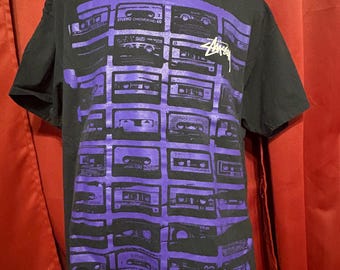 May include: Black t-shirt featuring a purple cassette tape design. The shirt has a crew neck and short sleeves. The Stussy logo is visible on the front. The text "STUDIO CHROMCKG 60" is printed on the shirt.