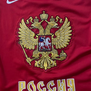 May include: Red and blue hockey jersey with a gold embroidered coat of arms featuring a double-headed eagle and the word "Россия" (Russia) in gold lettering.