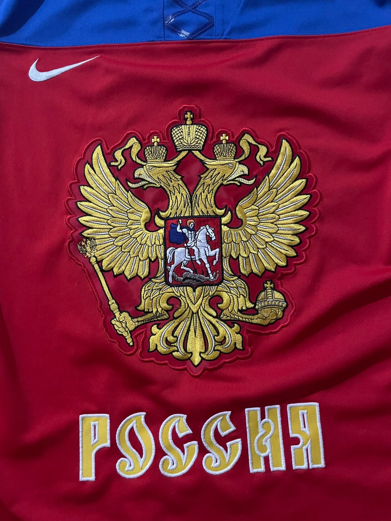 May include: Red and blue hockey jersey with a gold embroidered coat of arms featuring a double-headed eagle and the word "Россия" (Russia) in gold lettering.