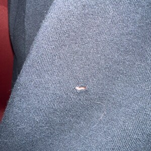May include: Close-up of a dark gray fabric with a small hole and a few stray fibers. The fabric appears to be a knit or woven material, possibly from a garment like a sweater or sweatshirt.