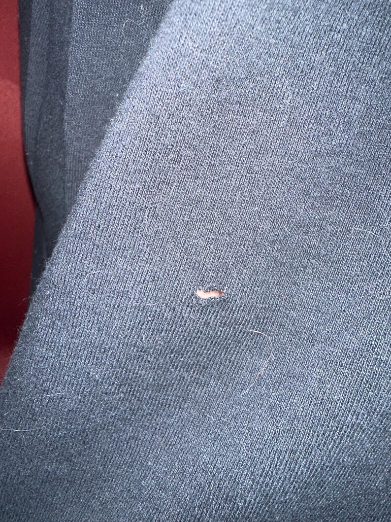 May include: Close-up of a dark gray fabric with a small hole and a few stray fibers. The fabric appears to be a knit or woven material, possibly from a garment like a sweater or sweatshirt.