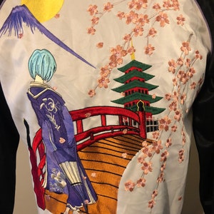 May include: A white satin bomber jacket with an embroidered design of a woman in a blue kimono walking across a red bridge. The bridge is over a river with a green pagoda in the background. The design also includes a mountain and a yellow moon. The jacket has black sleeves.