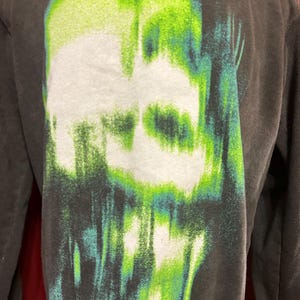 May include: Black long-sleeve shirt with a green and white graphic print of a skull. The skull is distorted and appears to be melting.