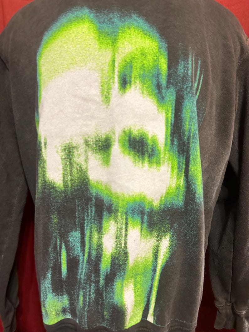 May include: Black long-sleeve shirt with a green and white graphic print of a skull. The skull is distorted and appears to be melting.