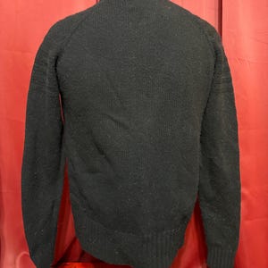 May include: A black sweater with a crew neck and long sleeves. The sweater is displayed on a mannequin against a red backdrop. The sleeves have ribbed detailing near the cuffs.