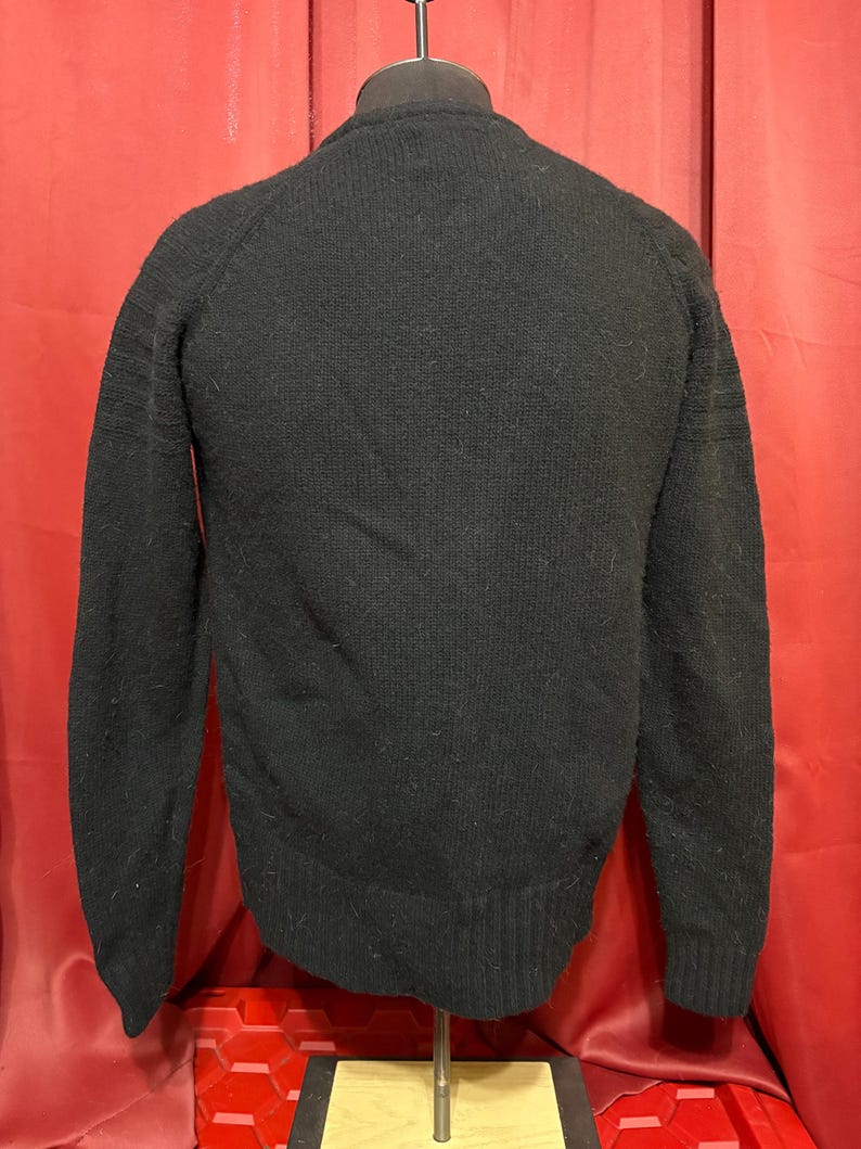 May include: A black sweater with a crew neck and long sleeves. The sweater is displayed on a mannequin against a red backdrop. The sleeves have ribbed detailing near the cuffs.