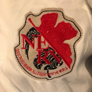 May include: A white patch with a black and red embroidered logo. The logo features a stylized leaf with a red center and black outline. The text "NERV" is embroidered in black with a black and white stylized leaf behind it. The text "GOD'S IN HIS HEAVEN ALL'S RIGHT WITH THE WORLD" is embroidered in black around the bottom of the logo.