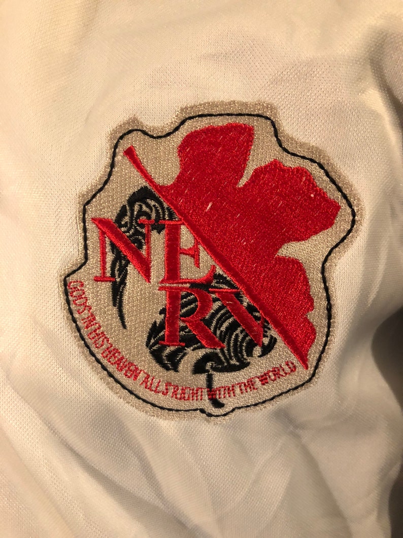 May include: A white patch with a black and red embroidered logo. The logo features a stylized leaf with a red center and black outline. The text "NERV" is embroidered in black with a black and white stylized leaf behind it. The text "GOD'S IN HIS HEAVEN ALL'S RIGHT WITH THE WORLD" is embroidered in black around the bottom of the logo.