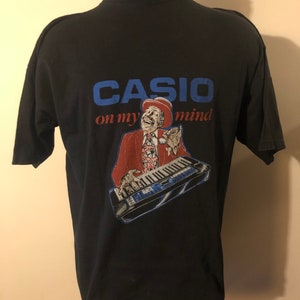 May include: Black t-shirt featuring a graphic of a man playing a keyboard. The graphic reads "CASIO on my mind".
