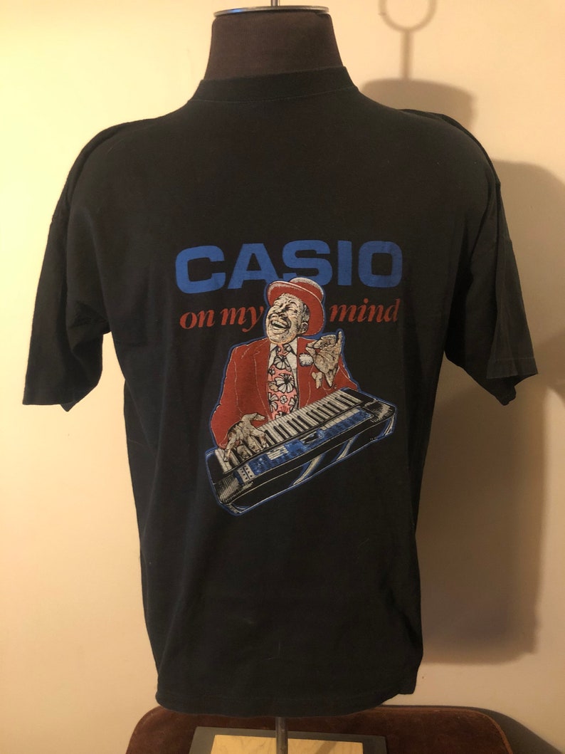 May include: Black t-shirt featuring a graphic of a man playing a keyboard. The graphic reads "CASIO on my mind".