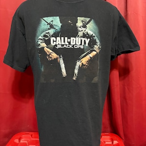 Puede incluir: Una camiseta negra de manga corta con un estampado gráfico de un soldado sosteniendo dos pistolas. El texto CALL OF DUTY BLACK OPS está impreso en blanco y amarillo sobre el soldado. El diseño incluye un fondo de color azul y verde oscuro.