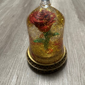 May include: A glass dome with a gold base encloses a single red rose with glitter. The rose is on a green stem and is surrounded by gold glitter.